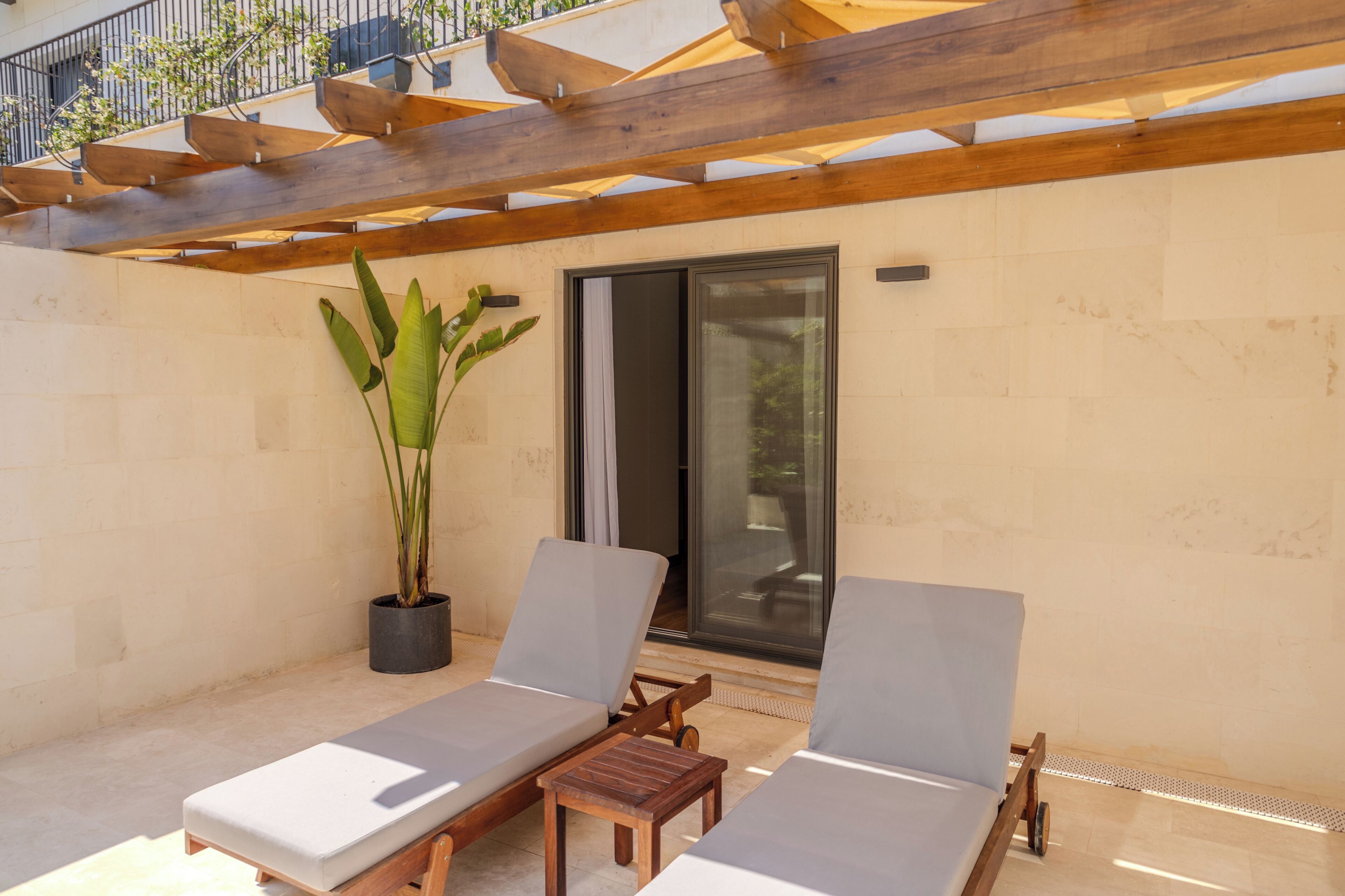 A covered outdoor seating area with two lounge chairs, a wooden pergola, a potted plant, and a sliding glass door.