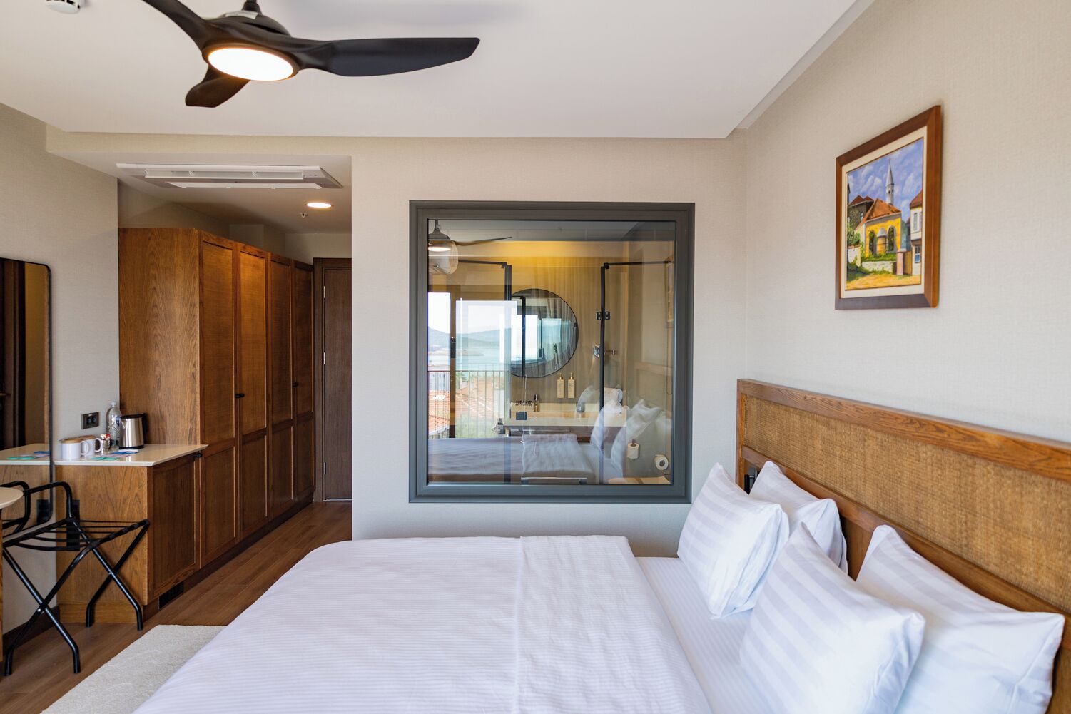 A hotel room with a large bed, a desk with a chair, a wardrobe, a ceiling fan, a large window with a view, and a framed picture on the wall.