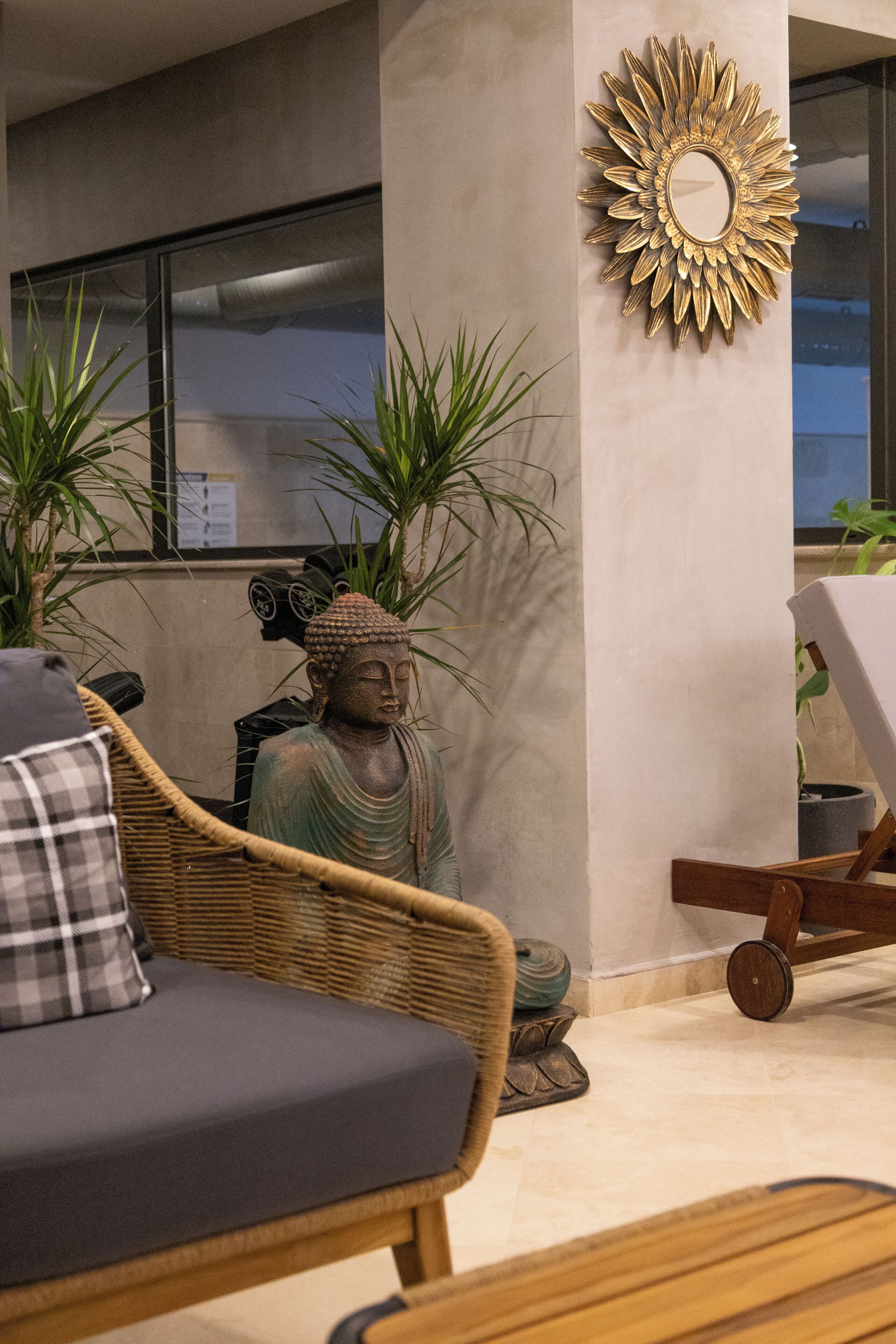 A room with a wicker sofa, a Buddha statue, a wall-mounted mirror, and potted plants.
