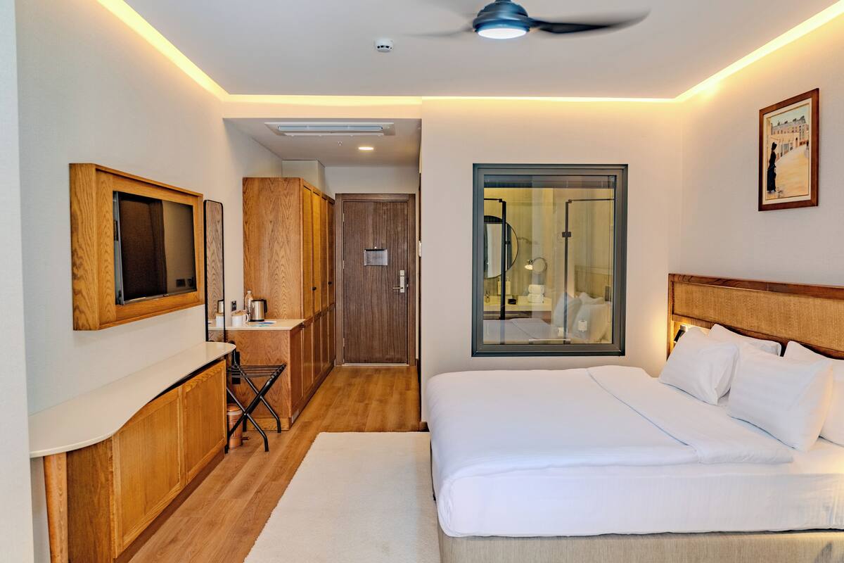 Ein Hotelzimmer mit Bett, Schreibtisch mit Stuhl, Spiegel, Fenster und Deckenventilator.