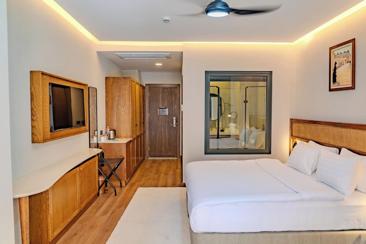 A hotel room with a bed, a desk with a chair, a mirror, a window, and a ceiling fan.