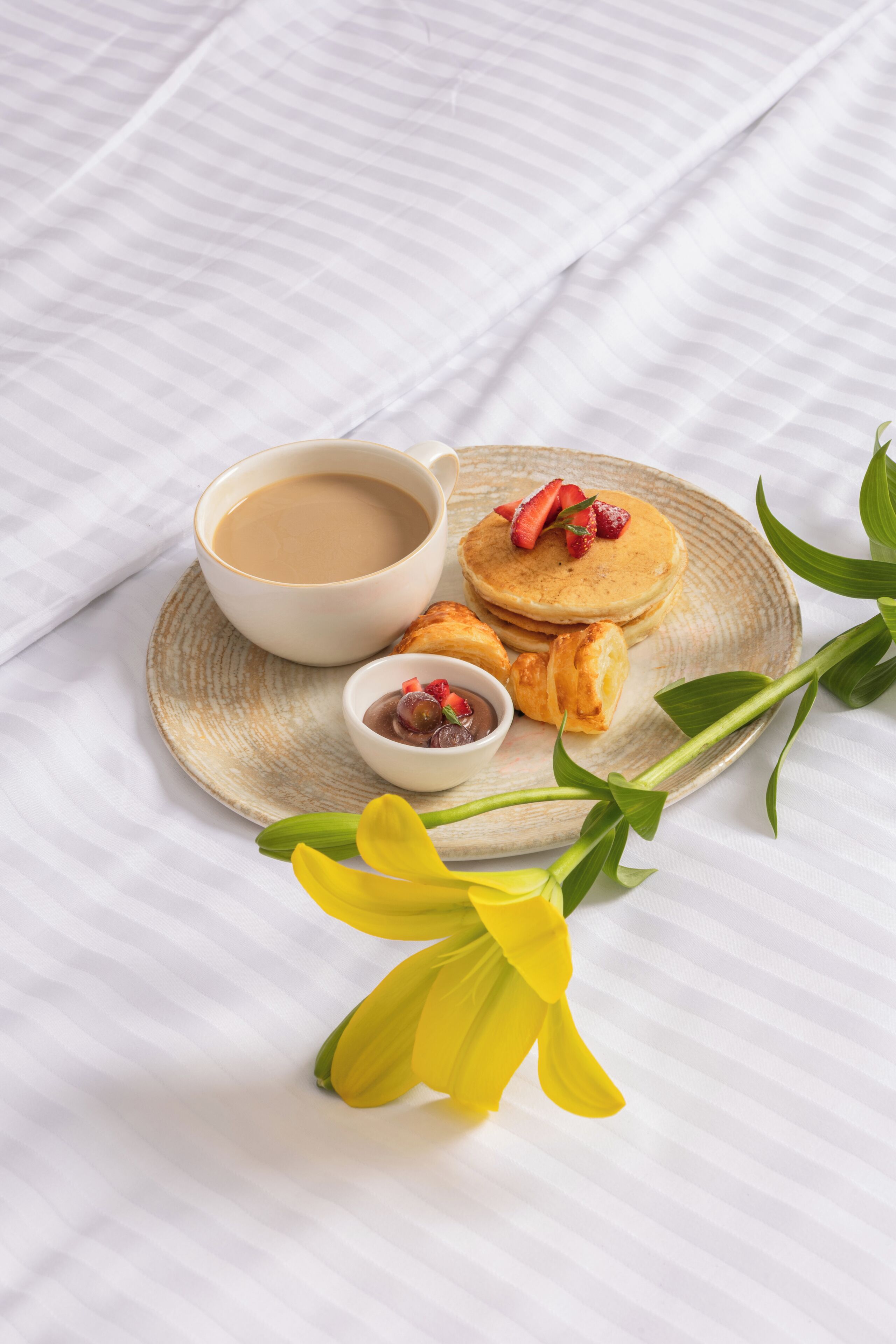 A breakfast setting with pancakes, a cup of coffee, and a small bowl of jam on a plate, accompanied by a yellow flower.