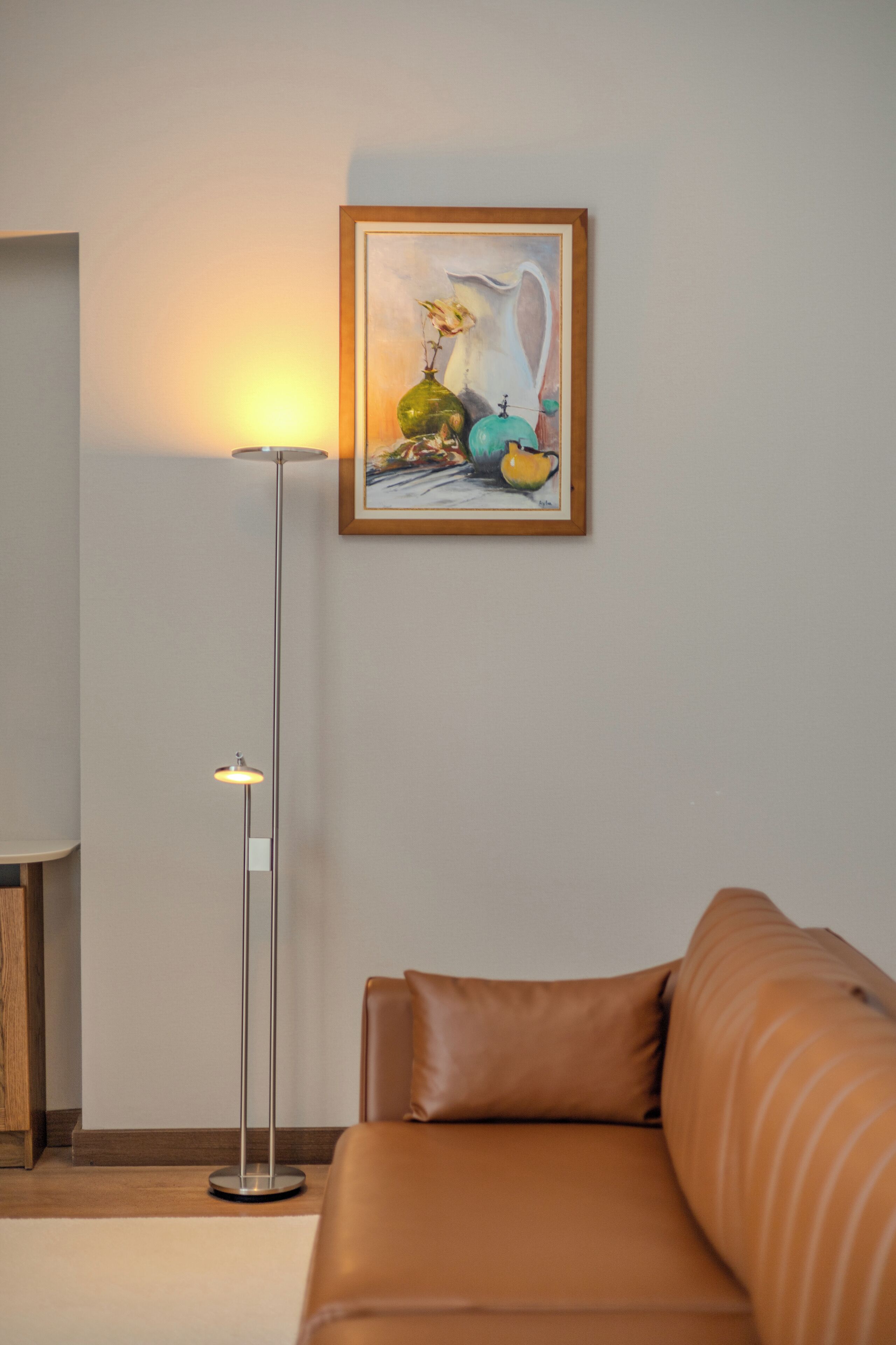 A room with a brown leather sofa, a floor lamp, and a framed artwork on the wall.