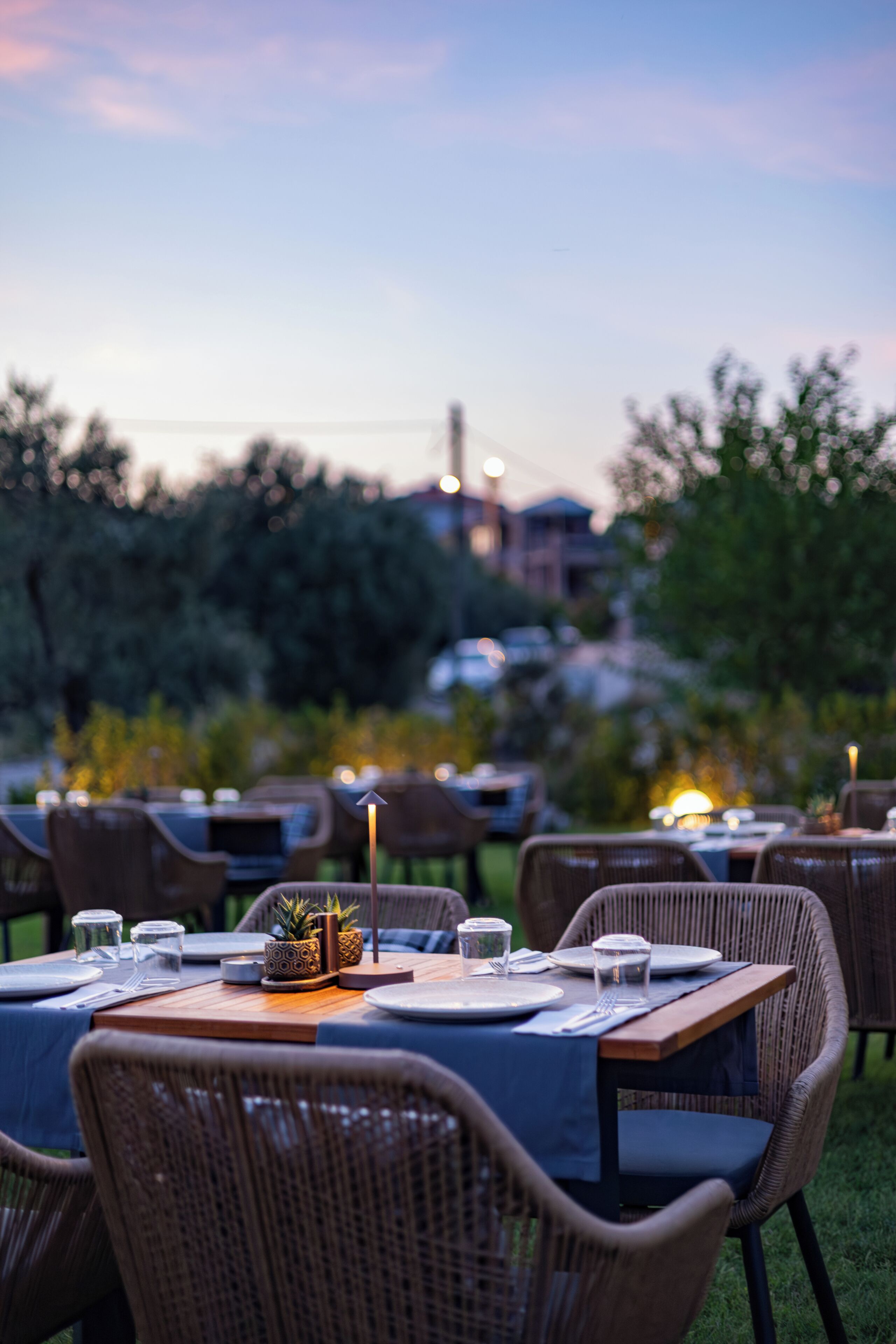 An outdoor dining area with wicker chairs and tables, set against a backdrop of trees and buildings.