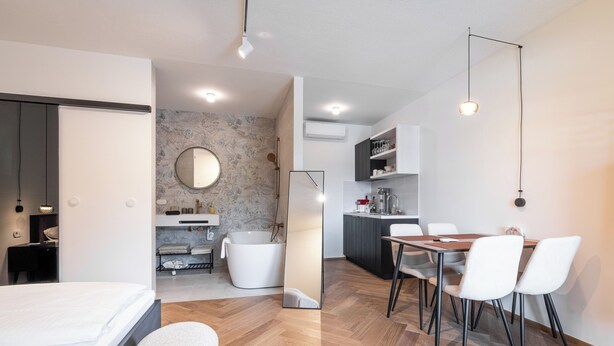 Una habitación de hotel moderna con suelo de madera, una cama, una zona de comedor con mesa y sillas, una cocina con armarios y un lavabo, y un baño con bañera y espejo.