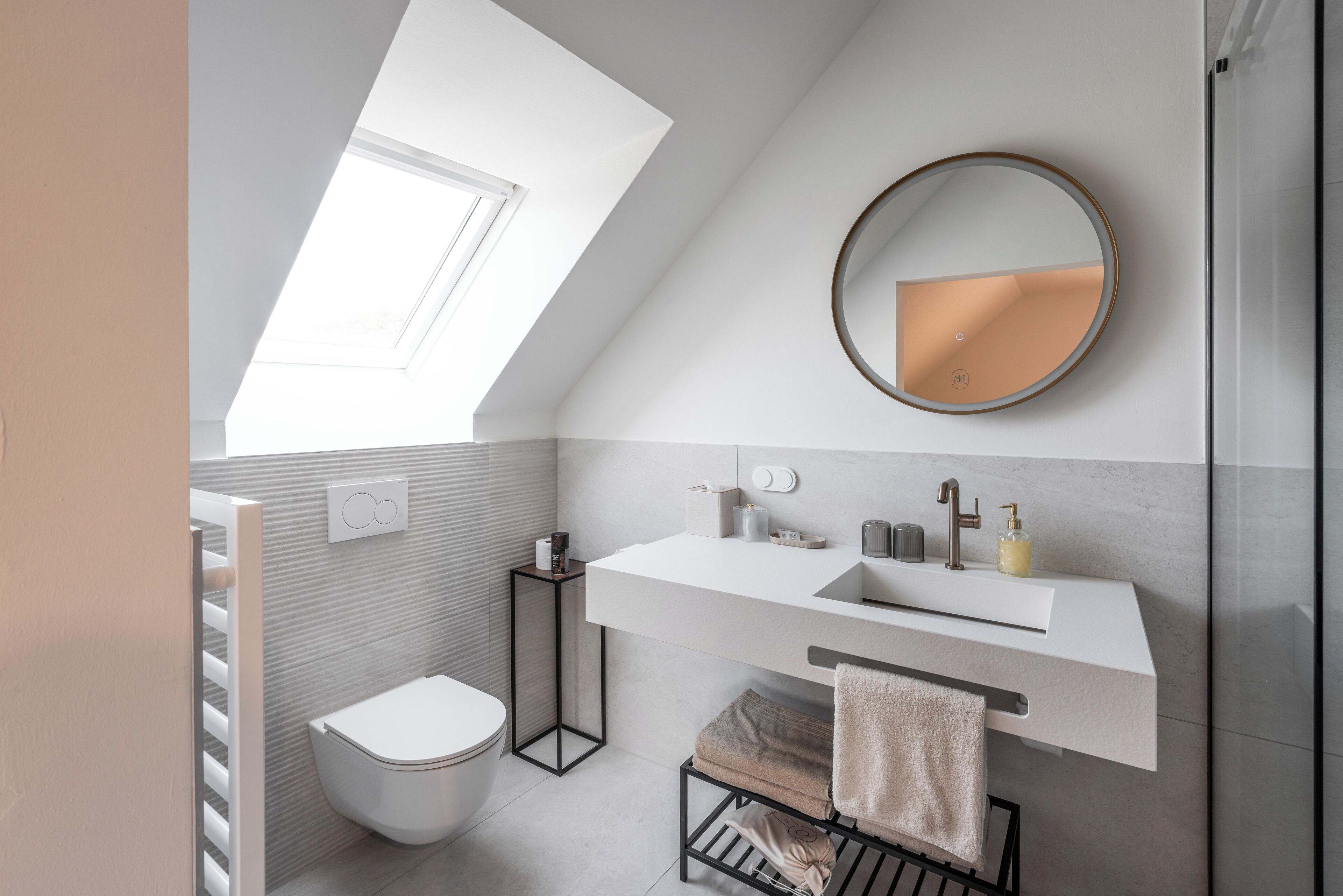 A modern bathroom with a skylight, a round mirror, a white sink, and a towel rack.
