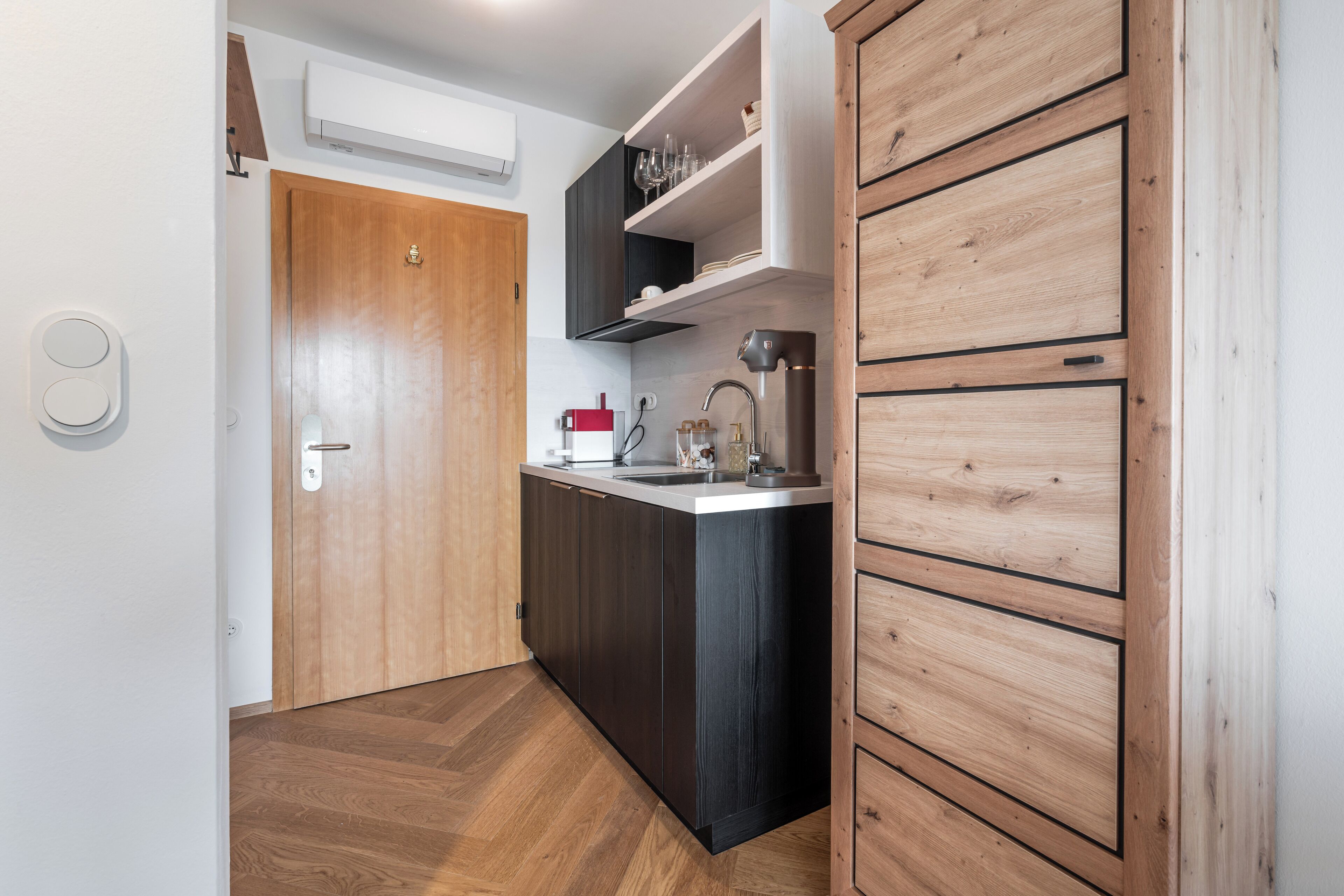 A compact kitchen with dark cabinetry, a wooden door, and a built-in shelf with glassware.