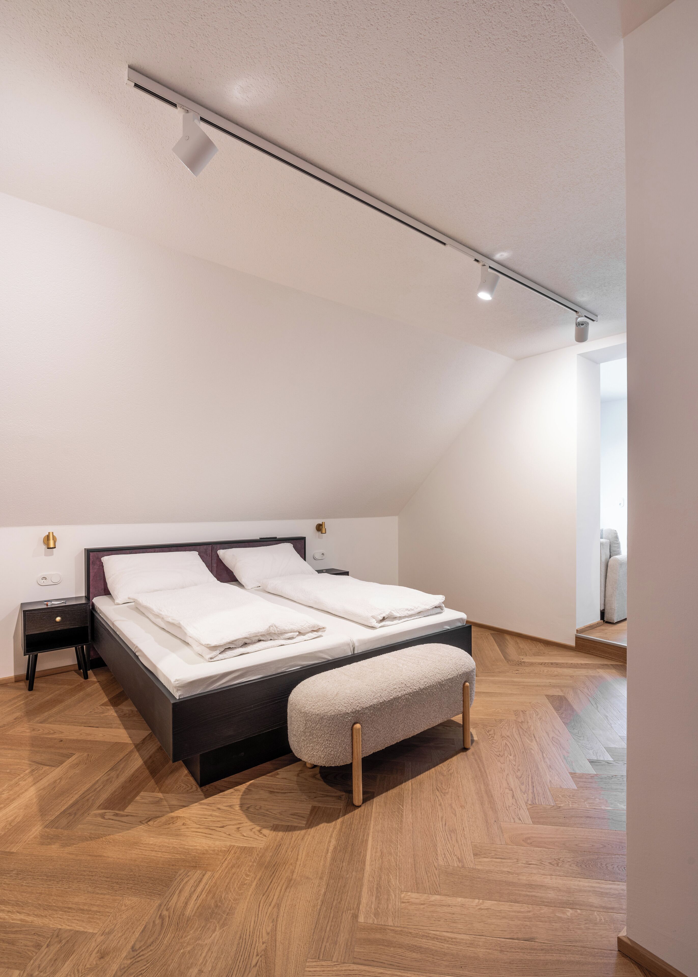 A modern bedroom with a wooden herringbone floor, a dark headboard, a white bed with multiple pillows, a bedside table, and a small bench.