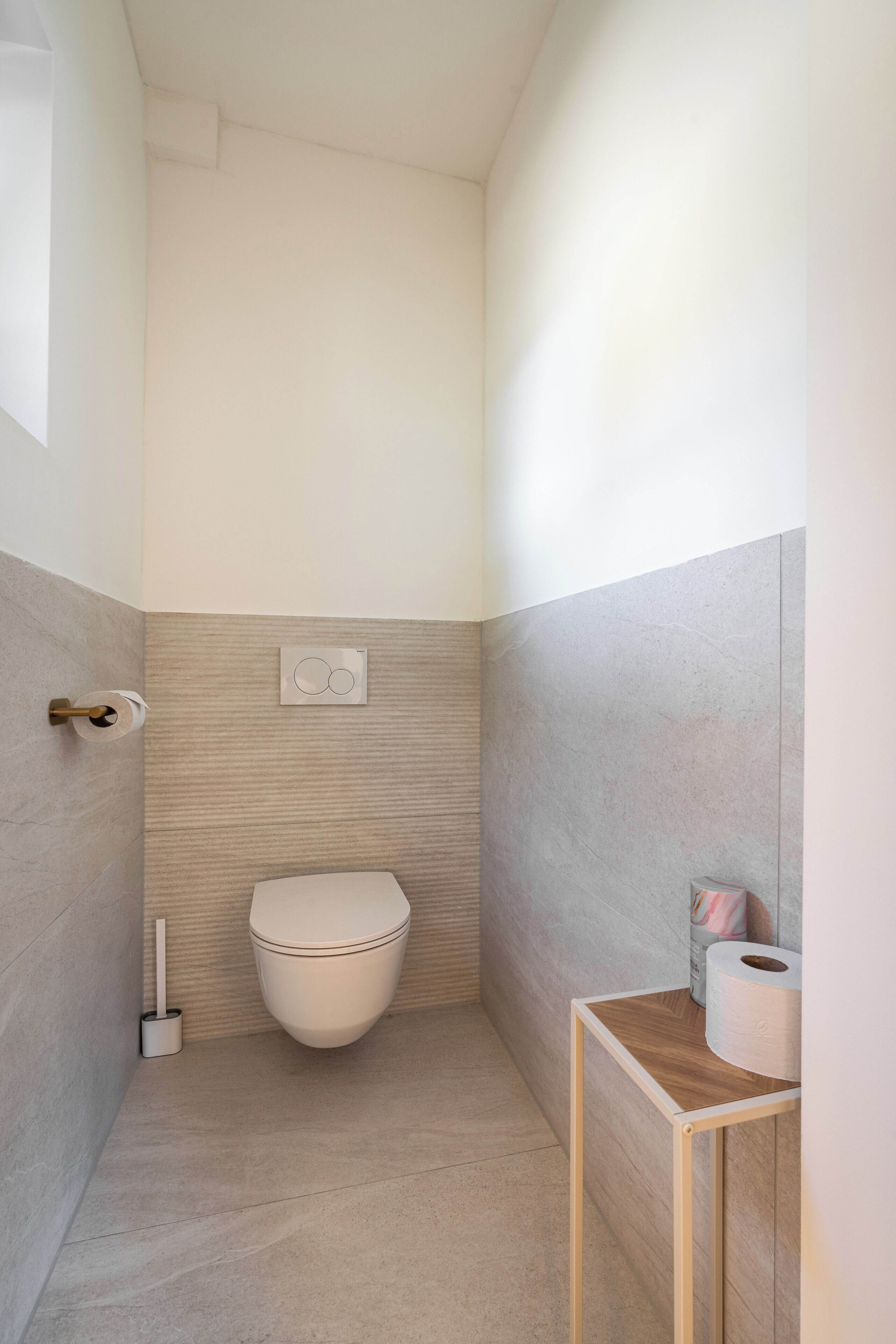 A compact bathroom with a toilet, a toilet paper holder, and a small wooden table with a roll of toilet paper.