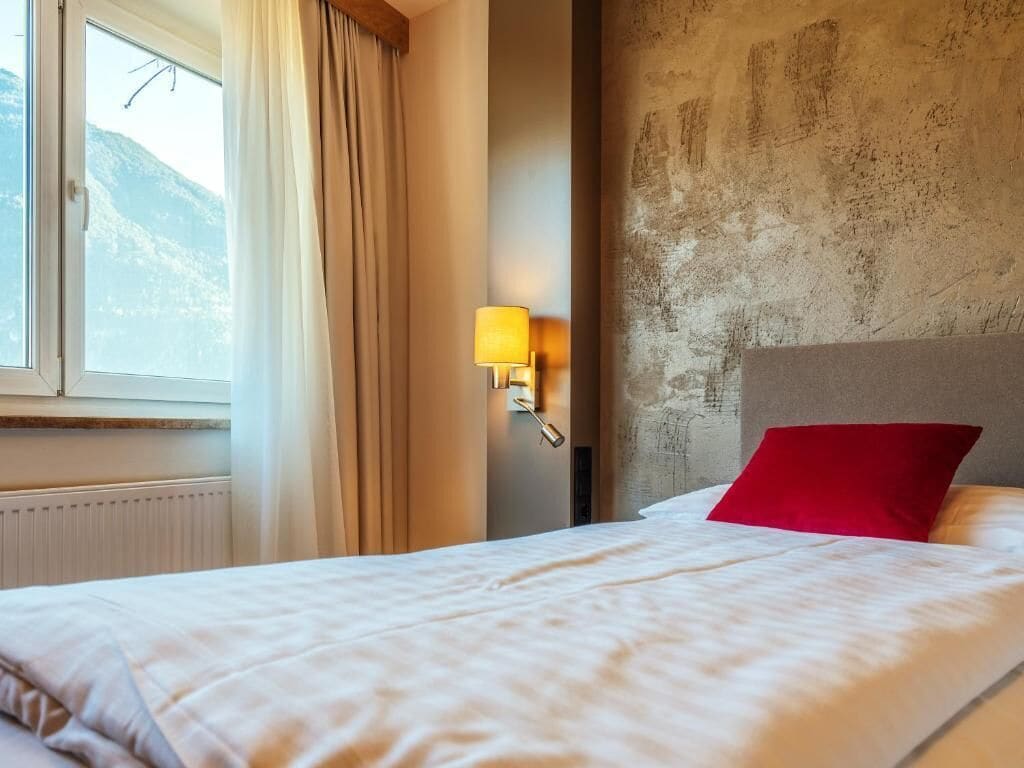 A hotel room with a bed, a window with curtains, a wall-mounted lamp, and a view of mountains.