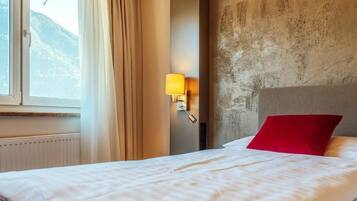 A hotel room with a bed, a window with curtains, a wall-mounted lamp, and a view of mountains.