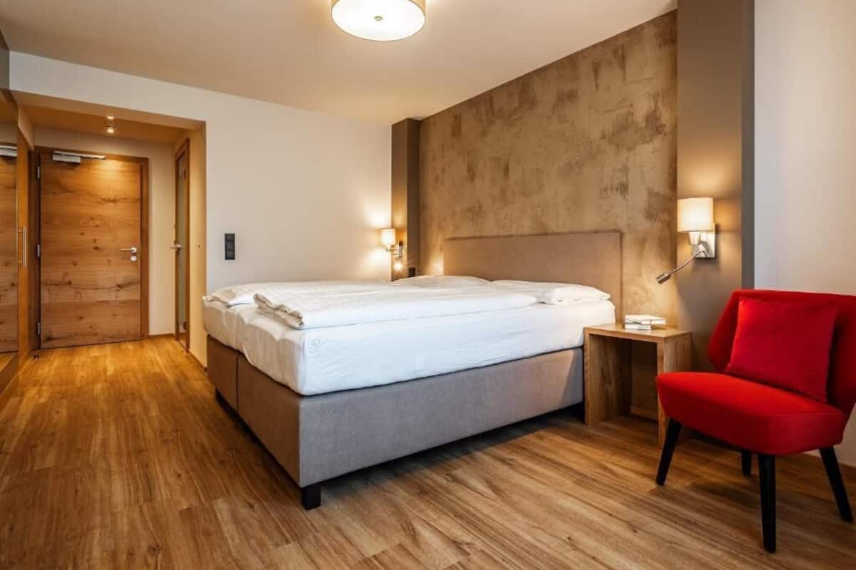 Una habitación de hotel moderna con piso de madera, una cama con sábanas blancas, una silla roja y una lámpara de pared.