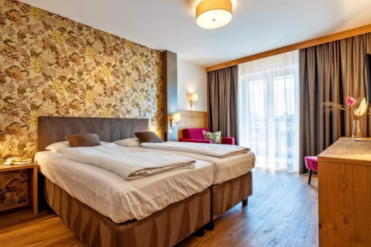 Habitación de hotel con dos camas, un escritorio, una silla, una lámpara y un ventanal grande con cortinas.