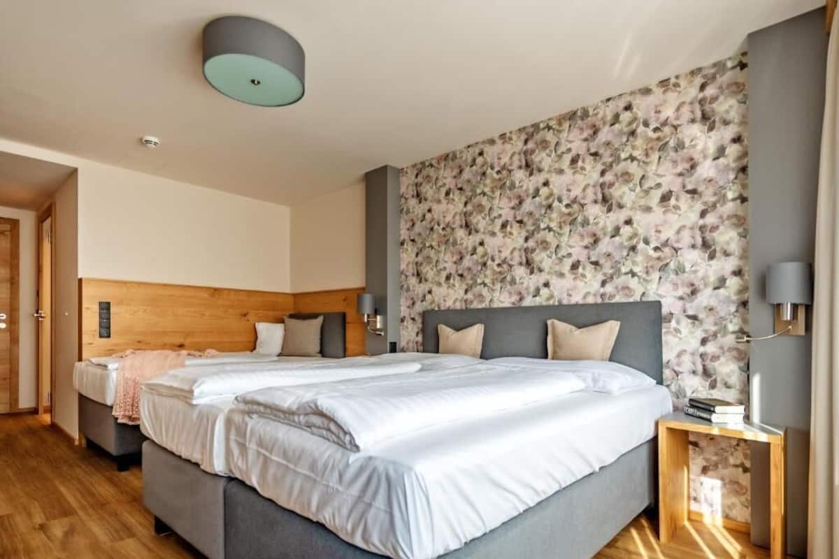 Habitación de hotel con una cama grande, cabecera de madera, papel tapiz floral y una mesita de noche con una lámpara.