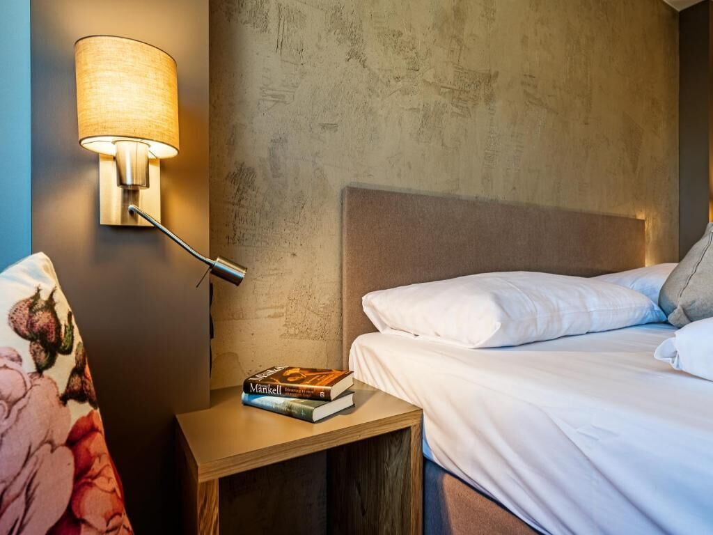 Un lit bien fait, avec du linge de lit blanc, une lampe de chevet et une petite table de nuit en bois sur laquelle repose un livre.