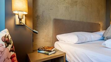 Una cama bien hecha con sábanas blancas, una lámpara de noche y una mesita de madera con un libro.