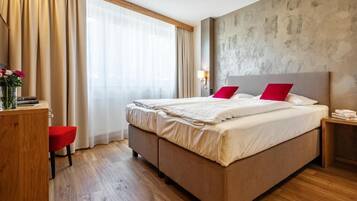 A double bed with red pillows, a wooden desk, a chair, and a bedside table with a lamp.