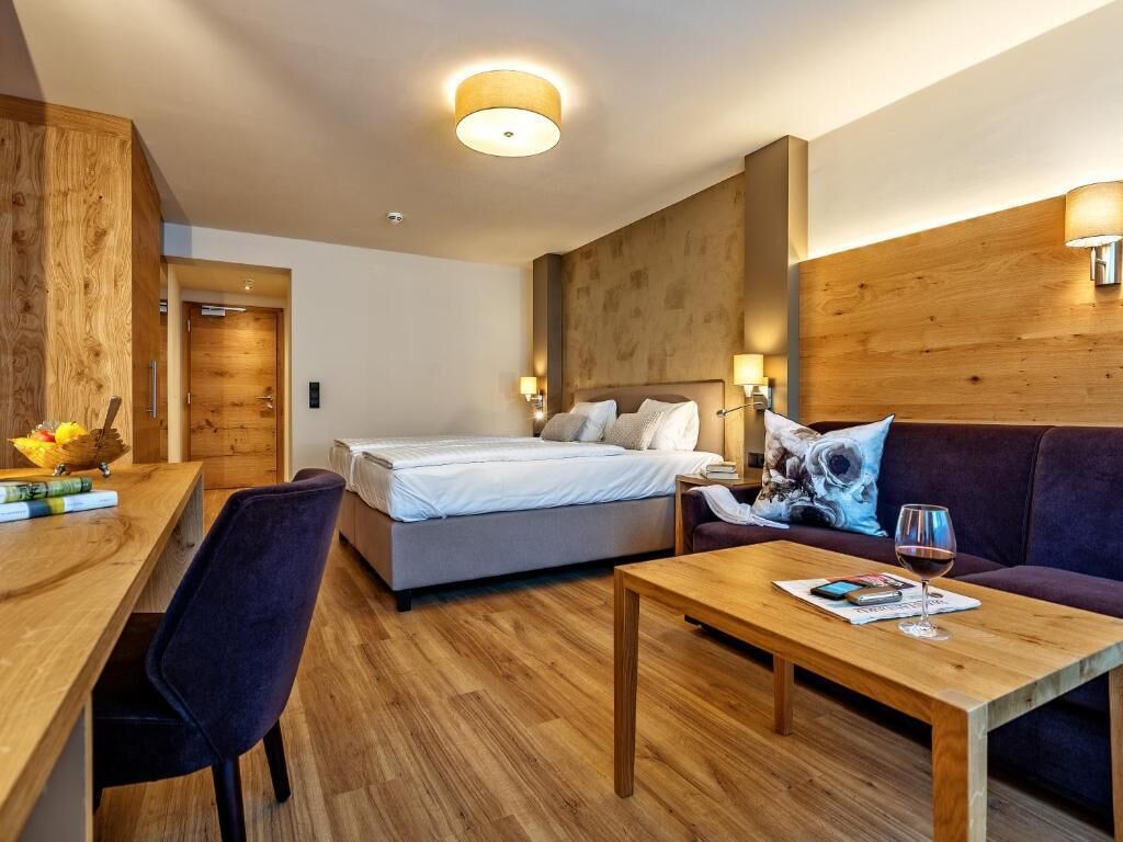 Una habitación de hotel moderna con una pared de acento de madera, una cama con ropa de cama blanca, un sofá, una mesa de centro de madera y un escritorio con una silla.