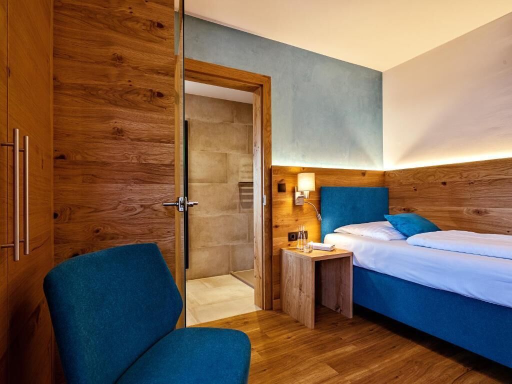 A room with a wooden wardrobe, a bed with blue bedding, a bedside table with a lamp, and a chair.