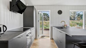 Une cuisine moderne dotée d’un grand îlot central, d’un four encastré et d’un évier avec un robinet doré. Un réveil est accroché au mur et une porte donne sur l’extérieur.