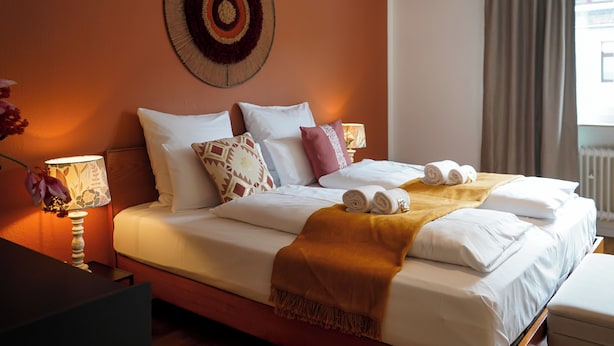 Una cama bien tendida con sábanas blancas y una manta marrón, un adorno en la pared y una ventana con cortinas.