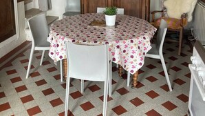 Een eetgedeelte met een ronde tafel, bedekt met een tafelkleed met bloemenprint, omringd door vier stoelen, een potplant en een houten kast. Daarboven hangt een plafondventilator.