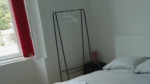 Ein ordentlich bezogenes Bett mit weißen Leinen und Handtüchern, ein Fenster mit roten Vorhängen und ein Wäscheständer an der Wand.