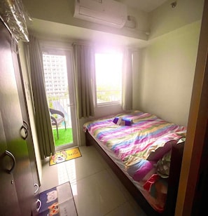 A small bedroom with a bed, a window, a door, and a view of a building outside.