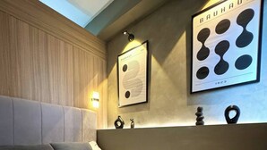 A modern hotel room with a large bed, a headboard, a wall-mounted artwork, a bedside table, and a basket with toiletries.