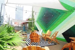 An outdoor seating area with wooden chairs and tables, a green wall mural, and string lights.