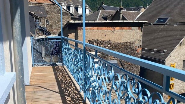 A balcony with a blue metal railing, wooden floor, and a view of buildings and mountains in the background.