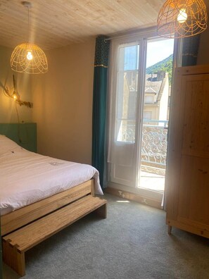 A bedroom with a bed, wooden headboard, two pendant lights, a sliding glass door, and a view of buildings outside.