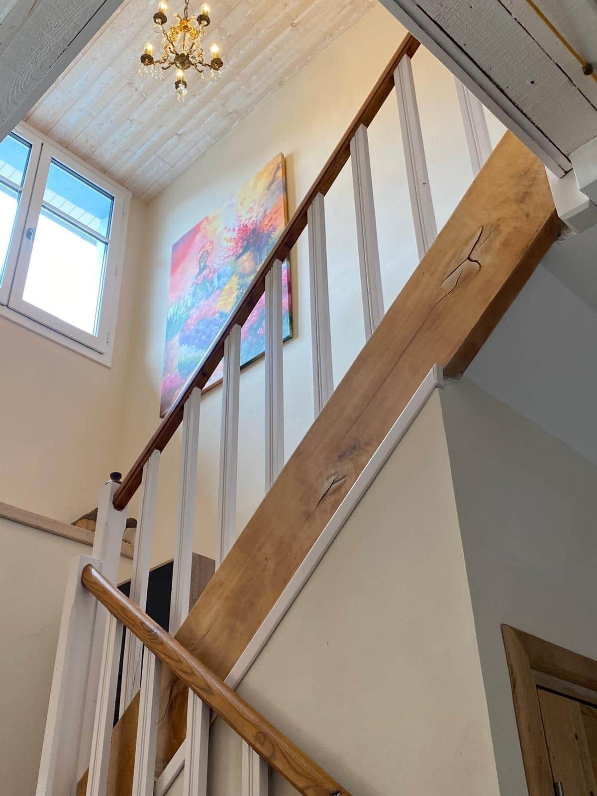 A staircase with wooden handrails and white walls, a chandelier, and a colorful painting on the wall.