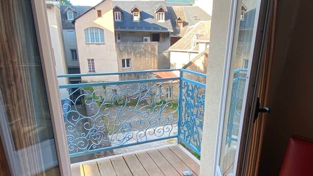 A balcony with a blue railing, a wooden floor, and a view of buildings with a tower. - Jasmine, pretty hat, on a bicycle (Bagnères-de-Luchon)