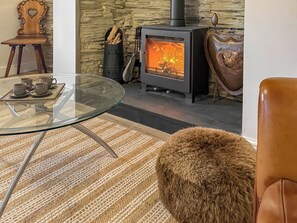 A cozy living room with a glass table, a wooden chair, a stone wall, a wood-burning stove, and a leather ottoman.