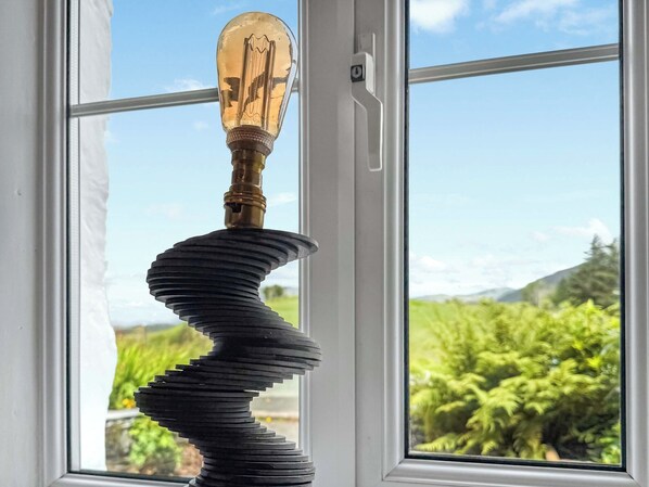 A spiral metal sculpture with a vintage light bulb on top, positioned in front of a window with a view of greenery and hills.