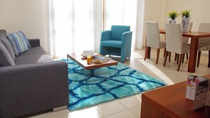 A modern living room with a grey sofa, a blue armchair, a wooden coffee table, and a dining area with a wooden table and chairs. - 1 Bedroom Apartment - BC11 (kouklia)