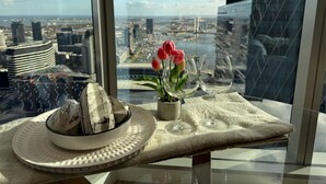 Un tavolo con un cappello, una ciotola, una pianta in vaso con fiori e bicchieri da vino su un tavolo di vetro che si affaccia sulla città.