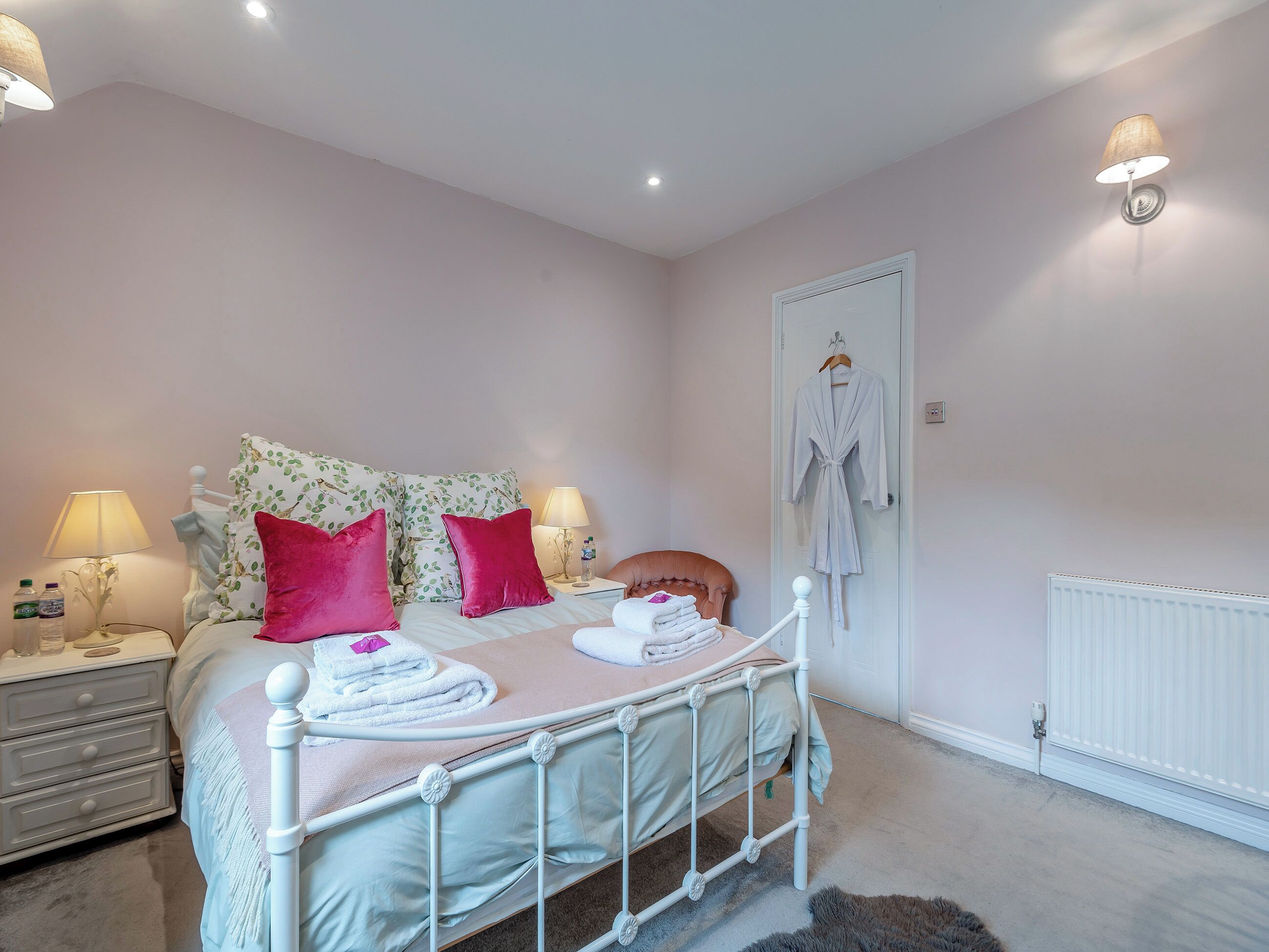 A neatly arranged bedroom with a bed, two bedside tables with lamps, a radiator, and a door with a robe hanging on it.