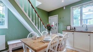 Un comedor con una mesa de madera y sillas blancas, una escalera con balaustres blancos, una cocina con armarios blancos y una ventana con vistas a follaje.