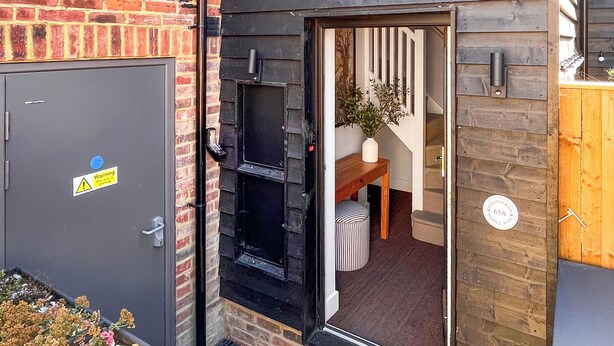 Une maison moderne avec une façade en briques, une porte grise ornée d’un panneau d’avertissement, un banc en bois, un pouf à rayures et une plante en pot.