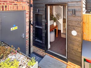 Une maison moderne avec une façade en briques, une porte grise ornée d’un panneau d’avertissement, un banc en bois, un pouf à rayures et une plante en pot.