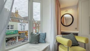 Une pièce avec un coin fenêtre, un fauteuil jaune, un miroir et une vue sur une rue commerçante.