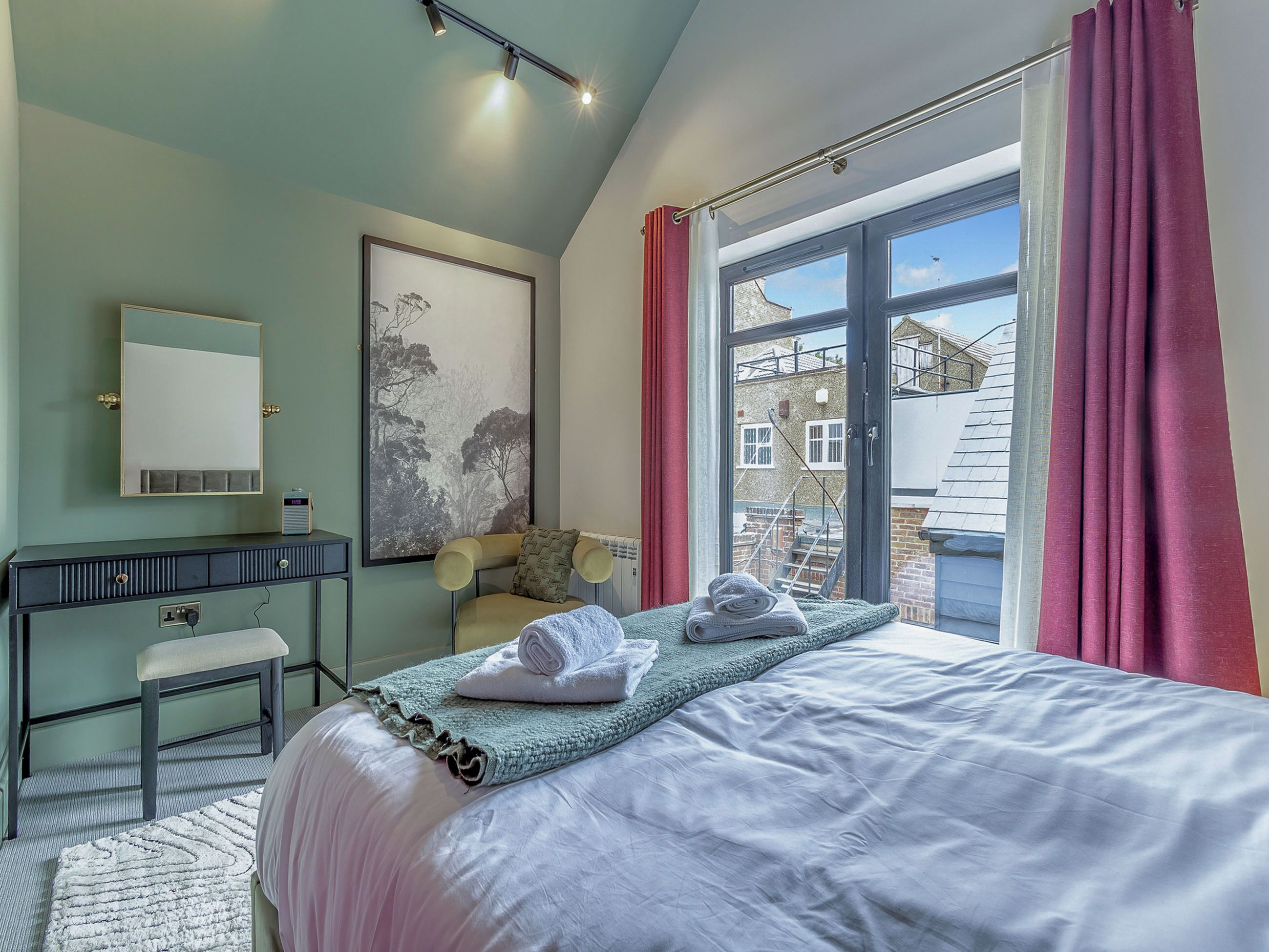 A bedroom with a bed, a desk, a chair, a window with curtains, and a view of buildings outside.