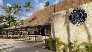 A stone building with a sign reading 'ES NIU,' outdoor seating with umbrellas, and palm trees.