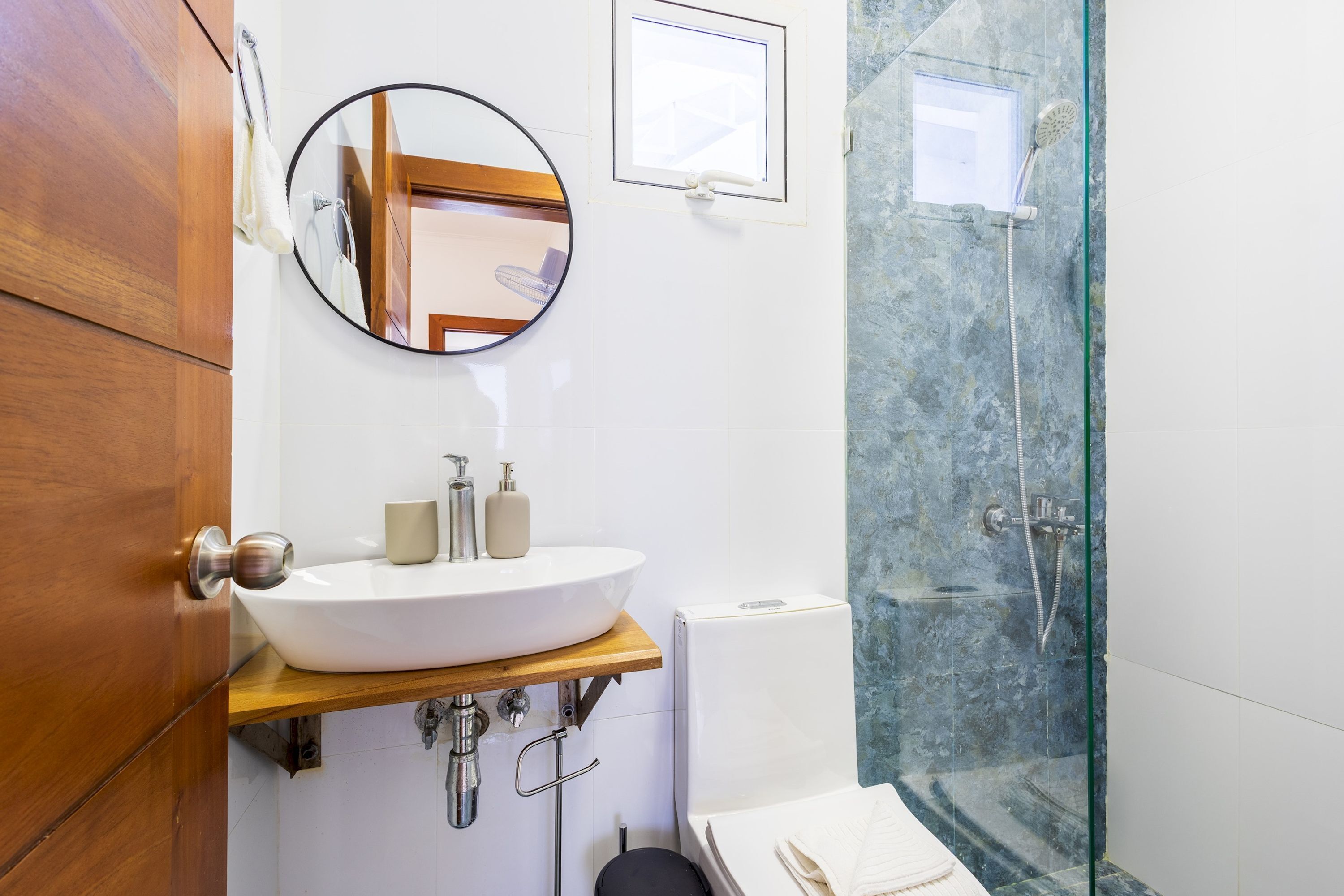 A bathroom with a wooden vanity, a round mirror, a white sink, a glass shower enclosure, and a towel on the floor.