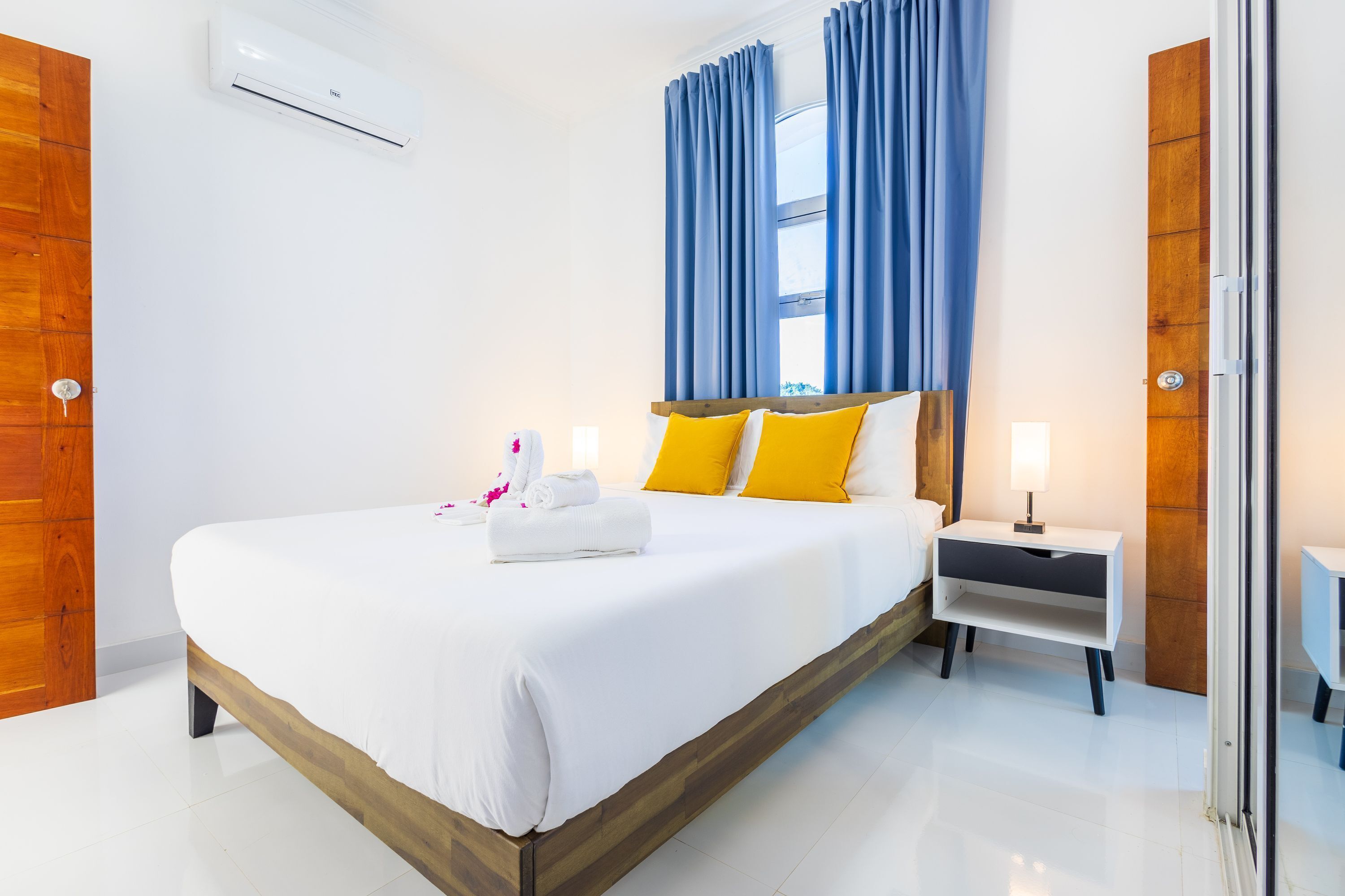 Una habitación de hotel moderna con una cama grande, cabecero de madera, ropa de cama blanca, una mesita de noche con lámpara y una ventana con cortinas azules.