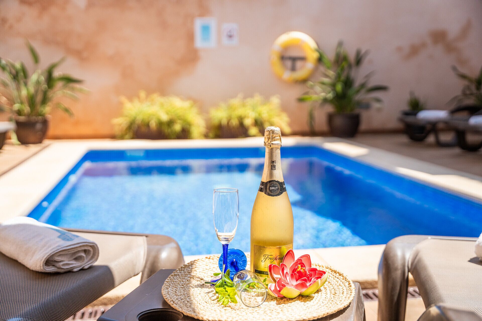 A poolside setting with a bottle of champagne, a glass, and decorative flowers on a woven mat.