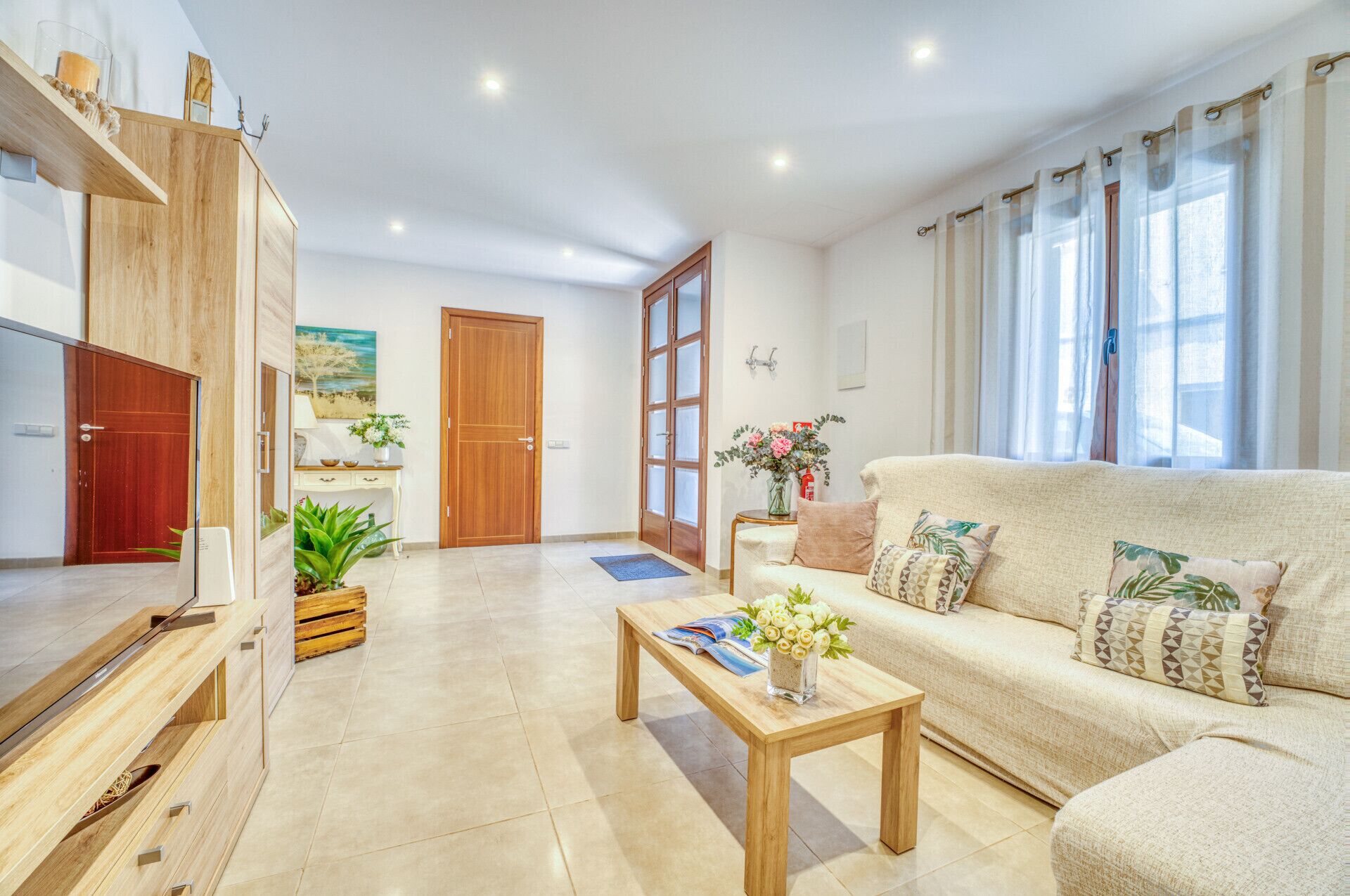 Un salon moderne avec un canapé beige, une table basse en bois et un meuble en bois. On y trouve une porte qui mène à une autre pièce, une petite table avec un vase de fleurs et un tableau accroché au mur.