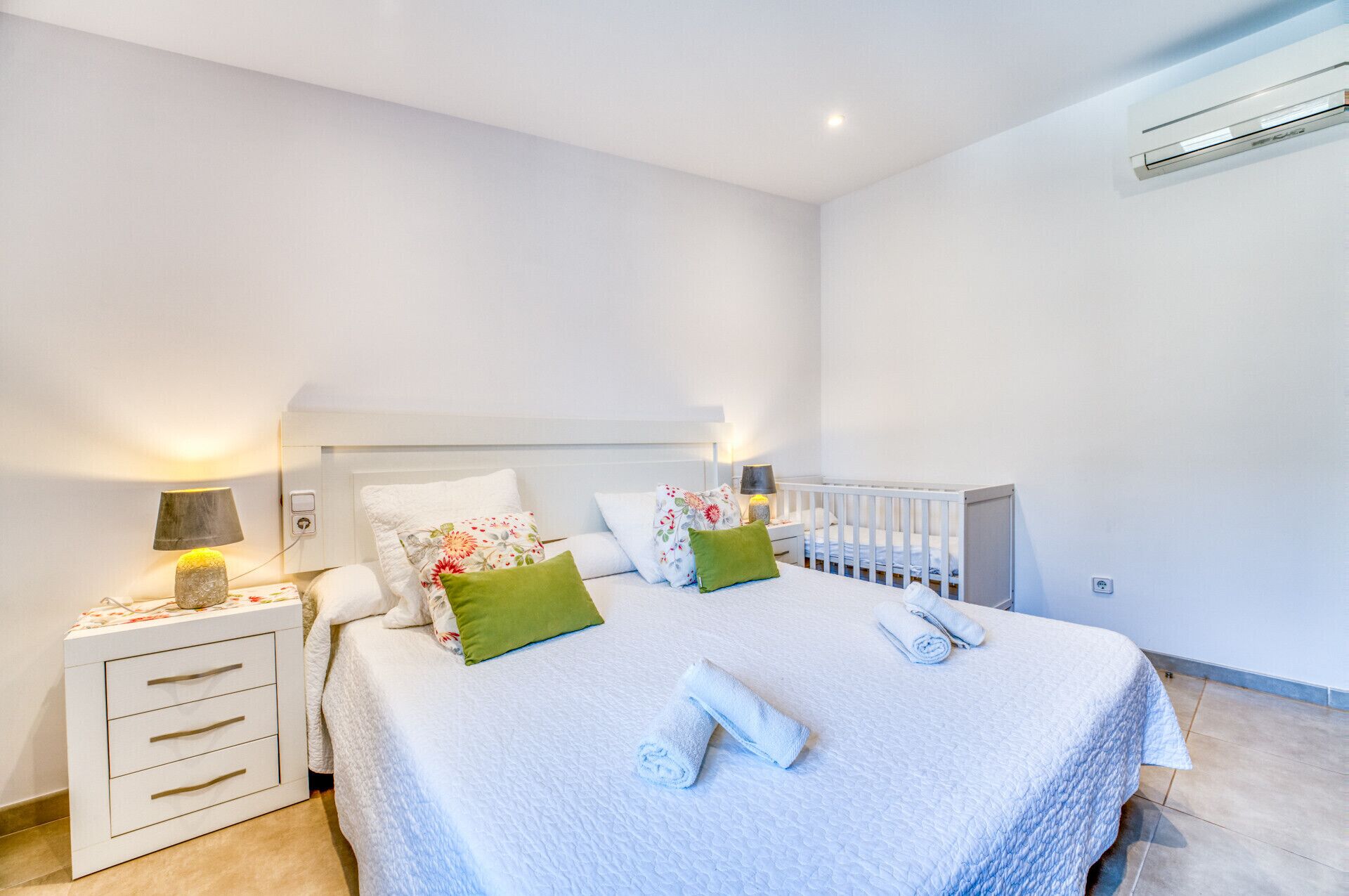 Une chambre à coucher comprenant un lit, une table de chevet, un berceau et un climatiseur fixé au mur.
